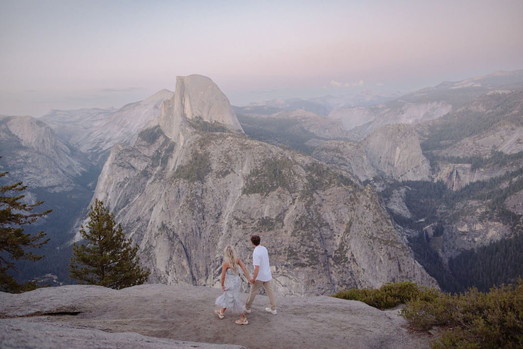 A couple stands under a large pine tree on a rocky overlook with mountains and Half Dome visible in the background for a yosemite national park proposal