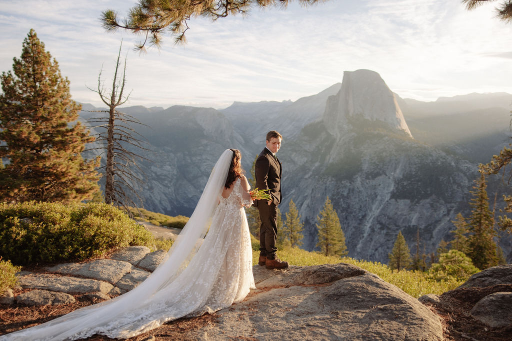 A bride and groom stand on rocky terrain overlooking a scenic mountain landscape at sunset, with the bride wearing a long white dress and veil for an elopement in Yosemite in September