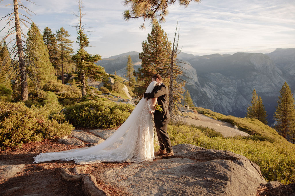 A bride and groom stand on rocky terrain overlooking a scenic mountain landscape at sunset, with the bride wearing a long white dress and veil for an elopement in Yosemite in September