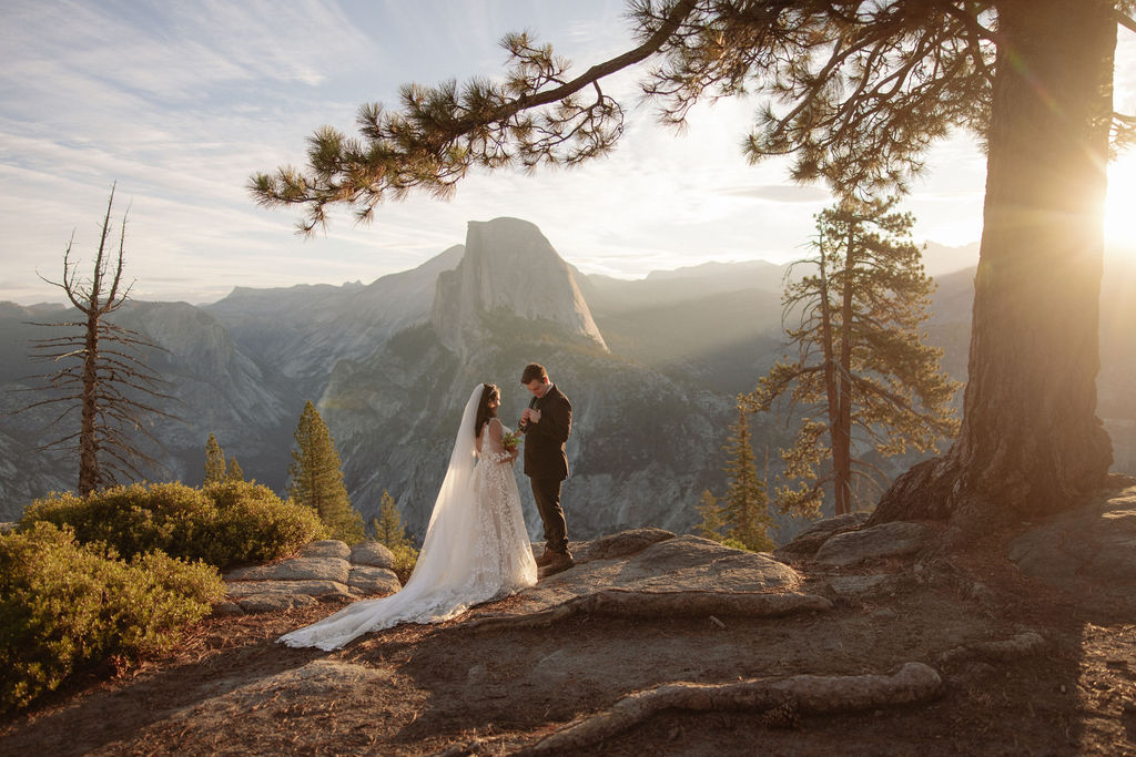 A bride and groom stand on rocky terrain overlooking a scenic mountain landscape at sunset, with the bride wearing a long white dress and veil for an elopement in Yosemite in September