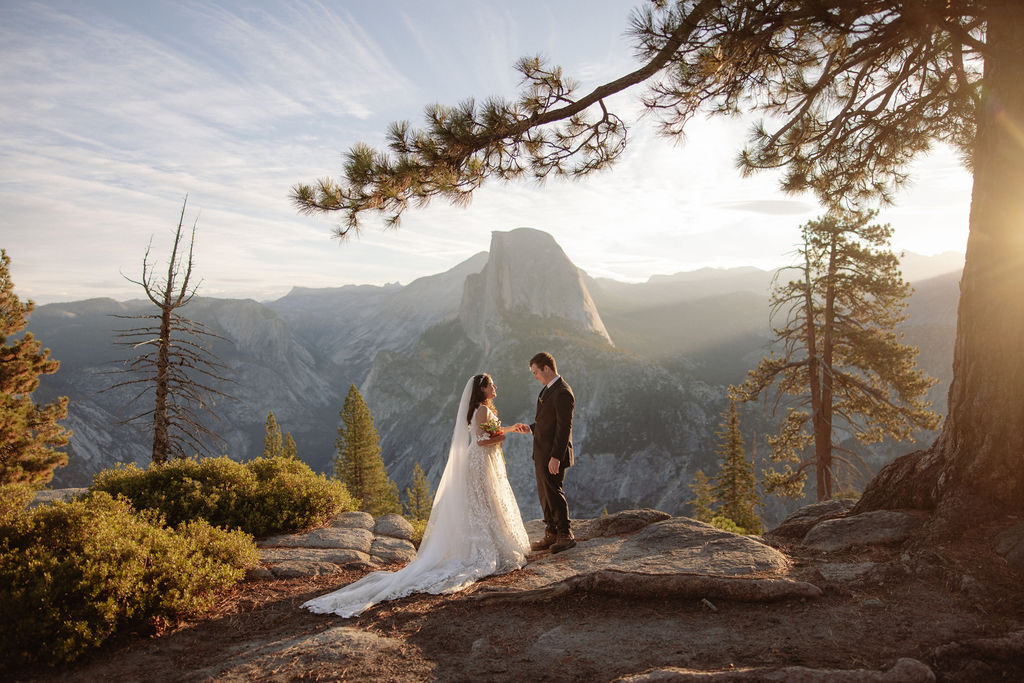 A bride and groom stand on rocky terrain overlooking a scenic mountain landscape at sunset, with the bride wearing a long white dress and veil for an elopement in Yosemite in September