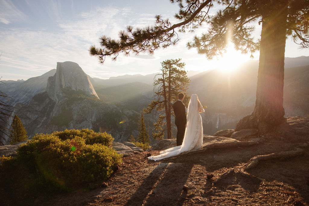 A bride and groom stand on rocky terrain overlooking a scenic mountain landscape at sunset, with the bride wearing a long white dress and veil for an elopement in Yosemite in September