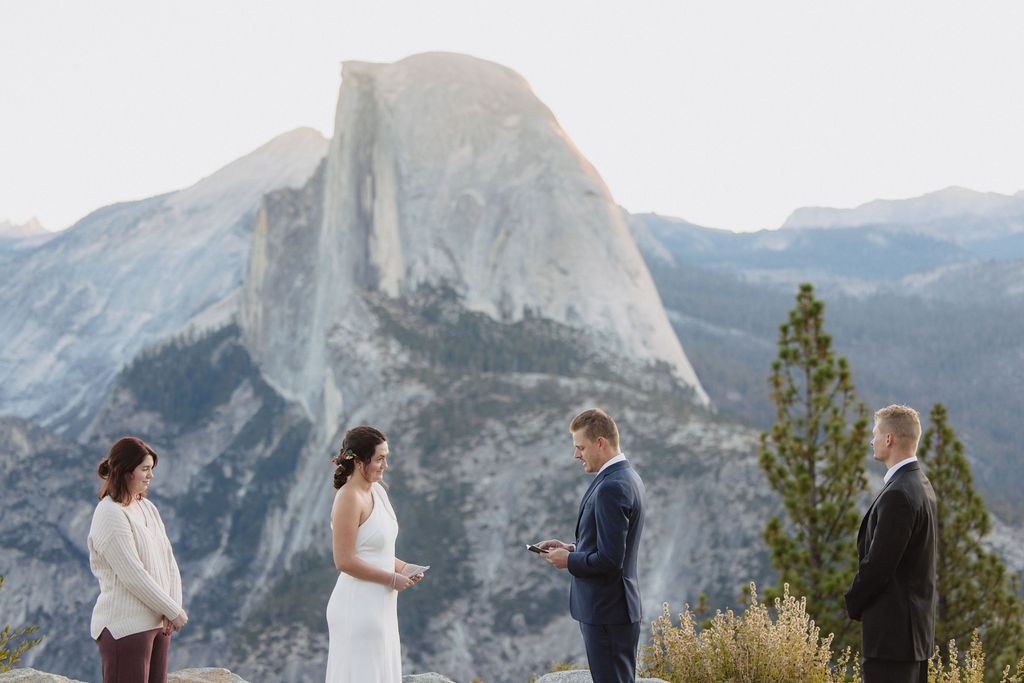 A couple exchanges vows outdoors with two witnesses, standing in front of a large mountain formation and pine trees in the background at Glacier Point Amphitheater
