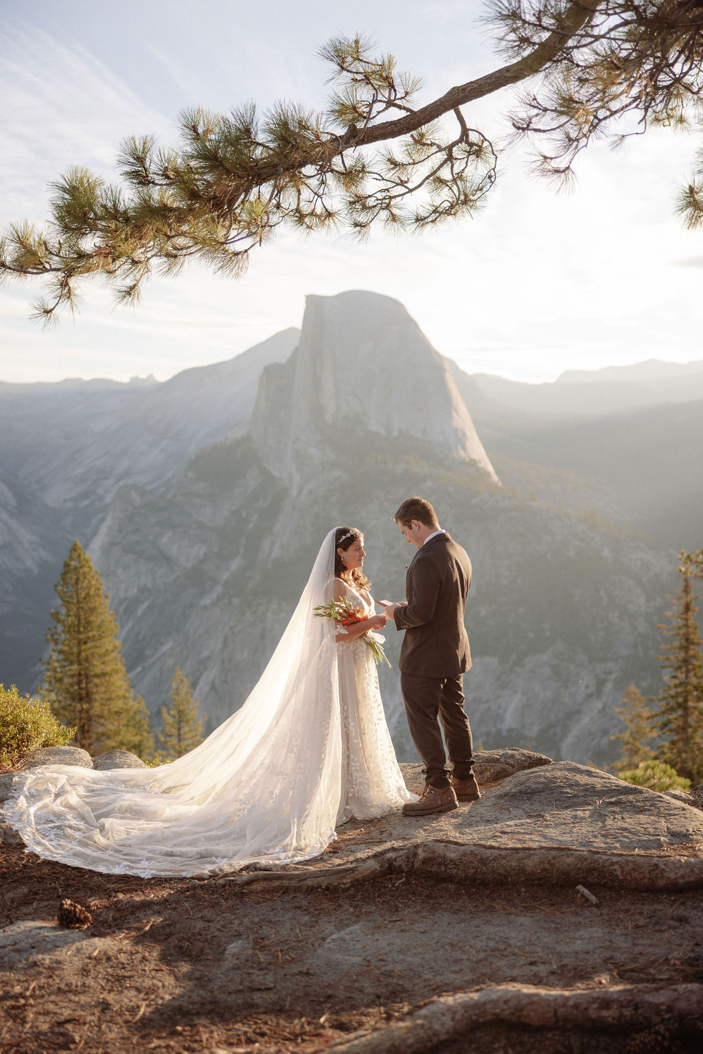 A bride and groom stand on rocky terrain overlooking a scenic mountain landscape at sunset, with the bride wearing a long white dress and veil for an elopement in Yosemite in September