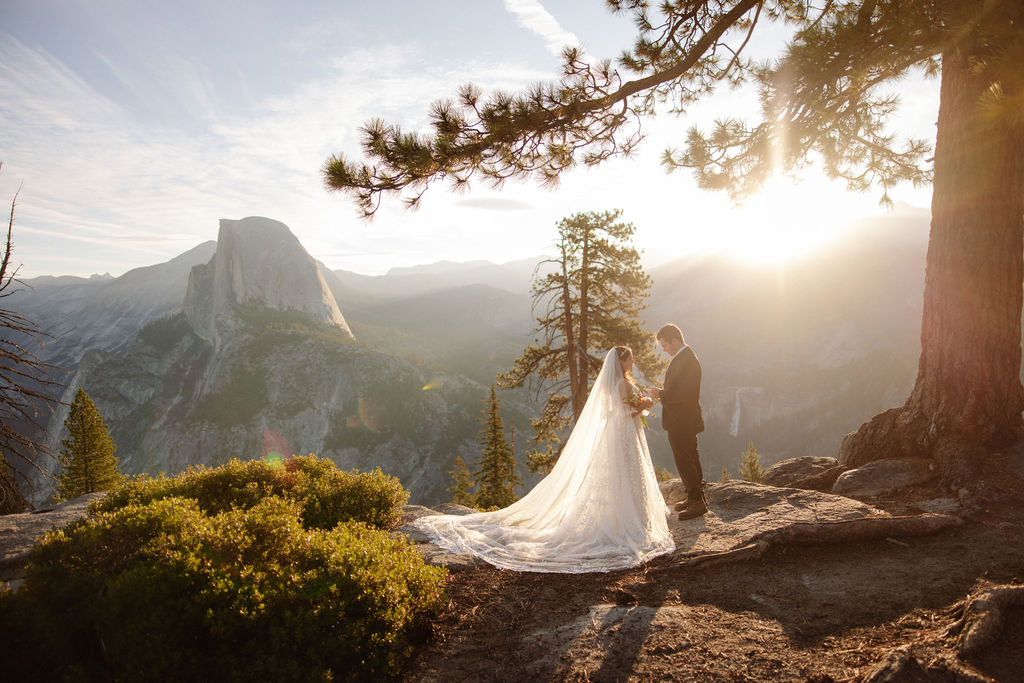 A bride and groom stand on rocky terrain overlooking a scenic mountain landscape at sunset, with the bride wearing a long white dress and veil for an elopement in Yosemite in September