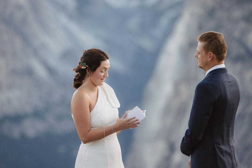 A couple exchanges vows outdoors with two witnesses, standing in front of a large mountain formation and pine trees in the background  at Glacier Point Amphitheater