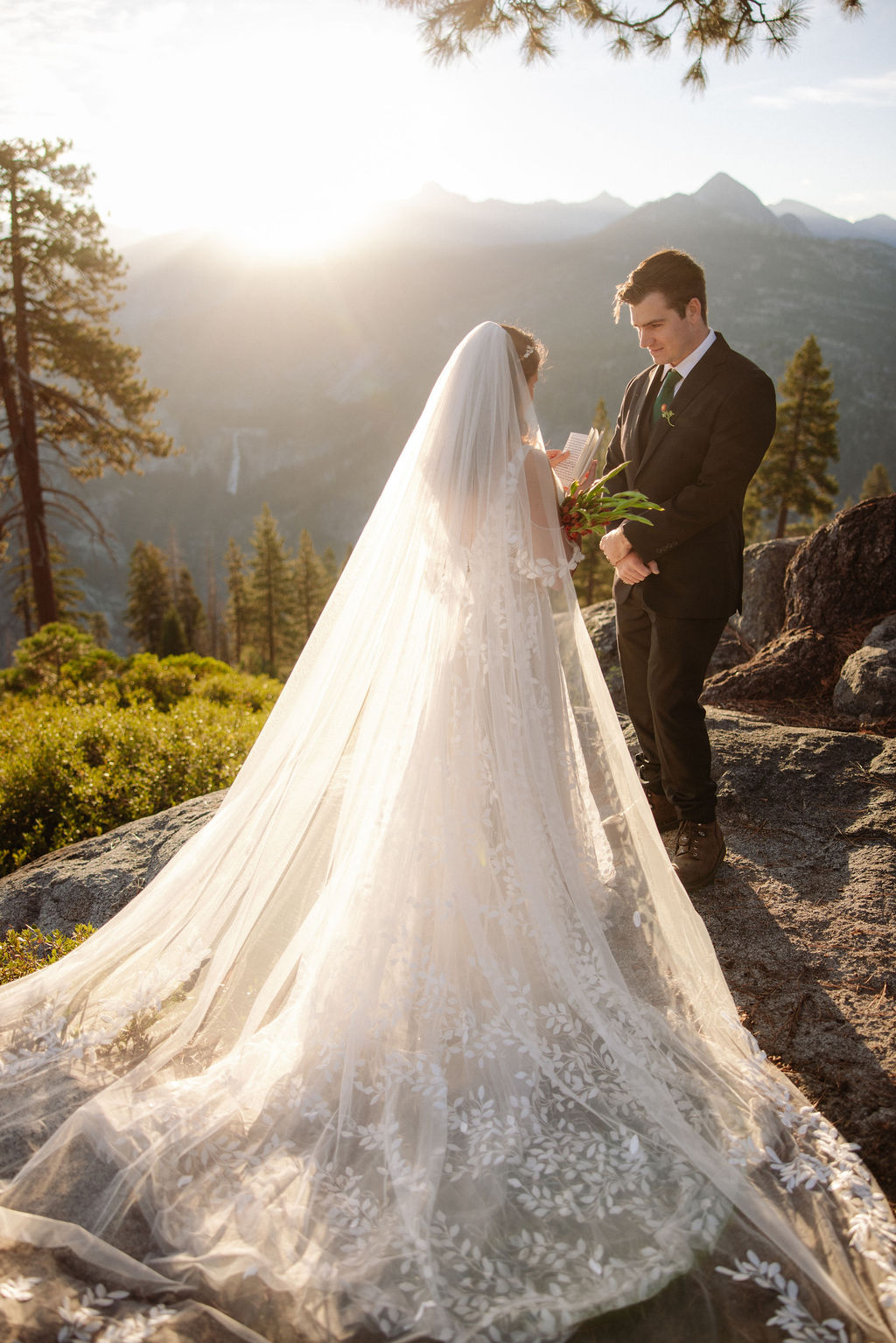 A bride and groom stand on rocky terrain overlooking a scenic mountain landscape at sunset, with the bride wearing a long white dress and veil for an elopement in Yosemite in September