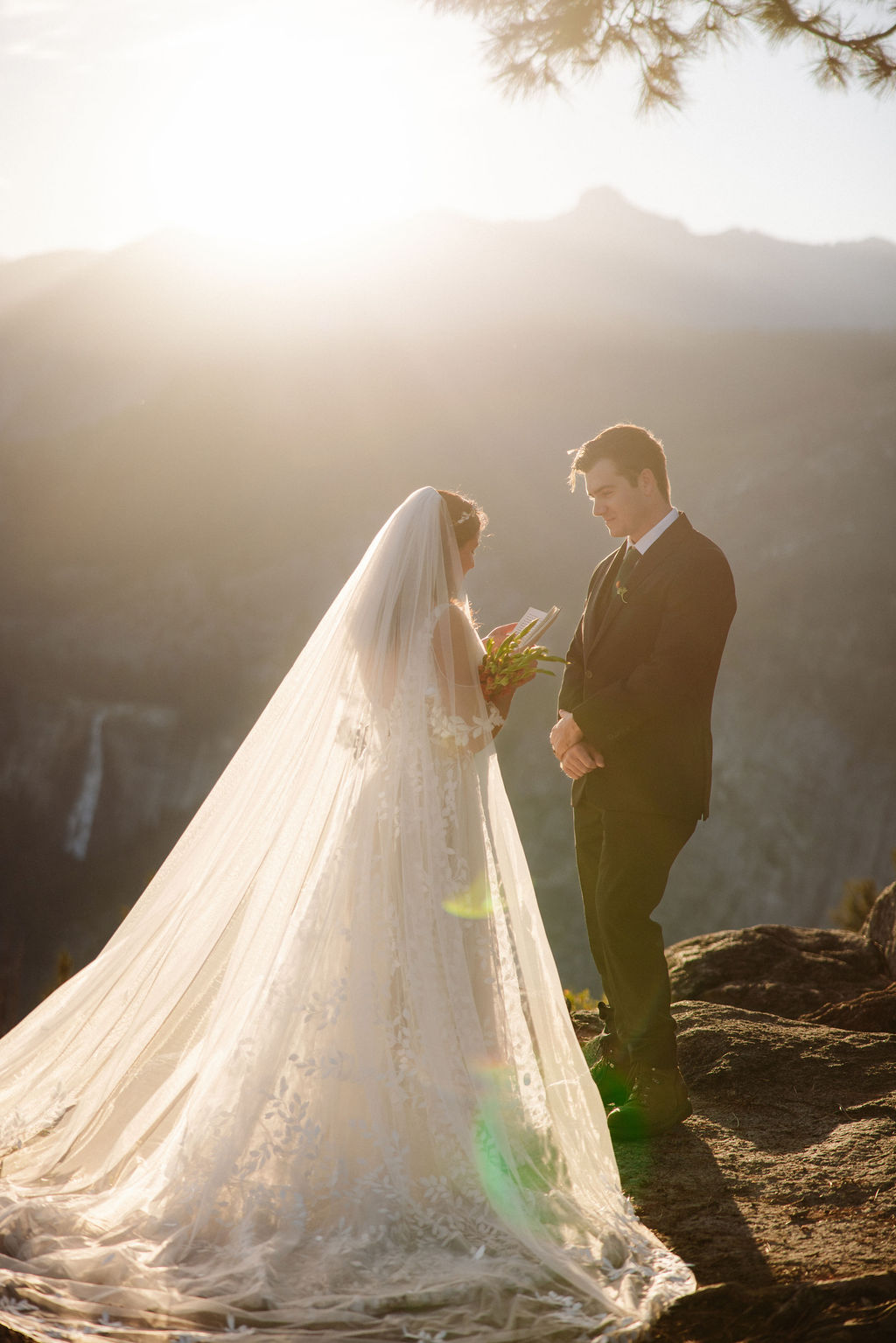 A bride and groom stand on rocky terrain overlooking a scenic mountain landscape at sunset, with the bride wearing a long white dress and veil for an elopement in Yosemite in September
