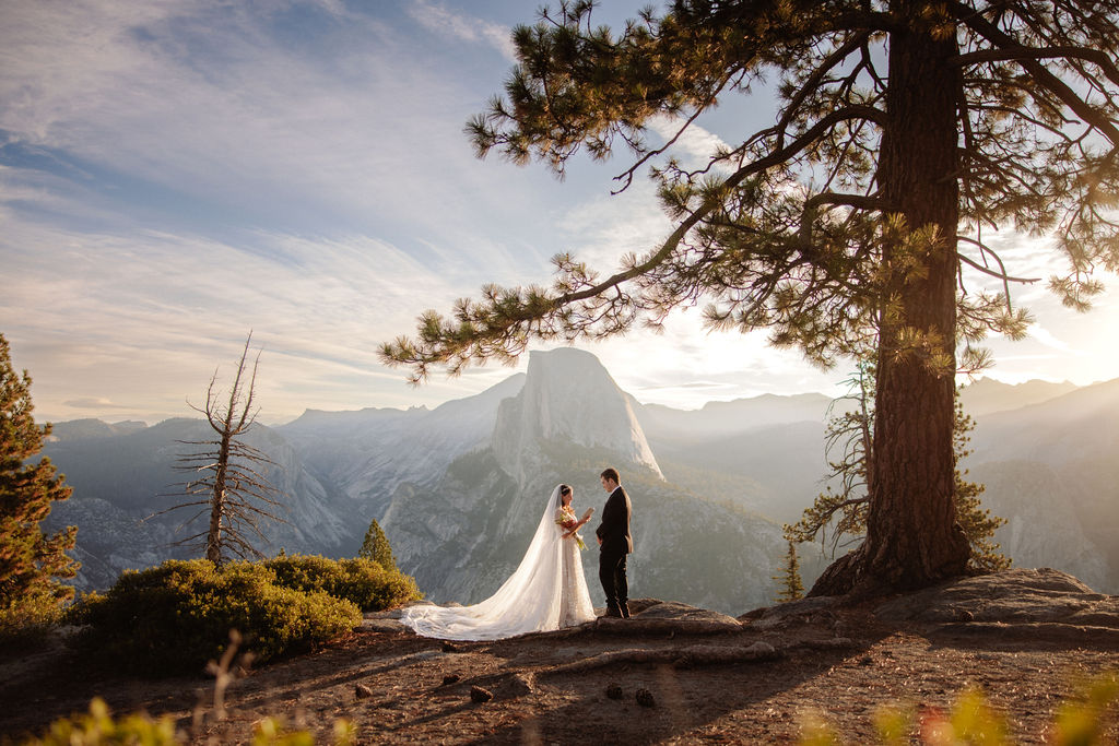 A bride and groom stand on rocky terrain overlooking a scenic mountain landscape at sunset, with the bride wearing a long white dress and veil for an elopement in Yosemite in September