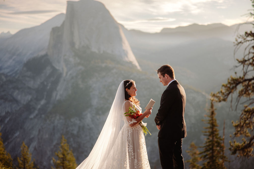 A bride and groom stand on rocky terrain overlooking a scenic mountain landscape at sunset, with the bride wearing a long white dress and veil for an elopement in Yosemite in September
