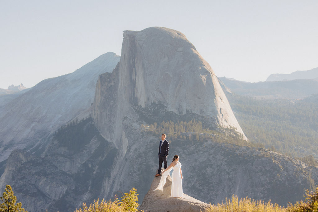 A bride and groom stand together on a rocky outcrop with mountain peaks, including Half Dome, visible in the background under a clear sky at Glacier Point Amphitheater
