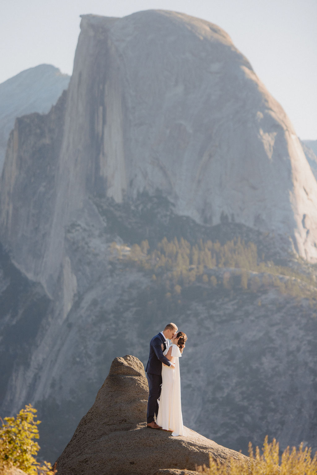 A bride and groom stand together on a rocky outcrop with mountain peaks, including Half Dome, visible in the background under a clear sky at Glacier Point Amphitheater