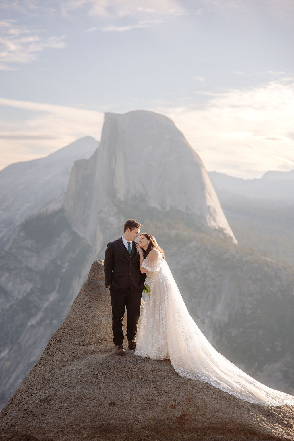 A bride and groom stand on rocky terrain overlooking a scenic mountain landscape at sunset, with the bride wearing a long white dress and veil for an elopement in Yosemite in September