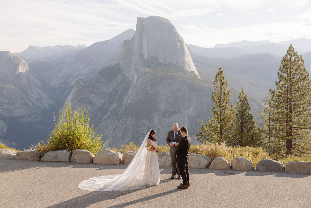 Bride and groom hold hands during an outdoor wedding ceremony with an officiant, set against a mountainous backdrop.