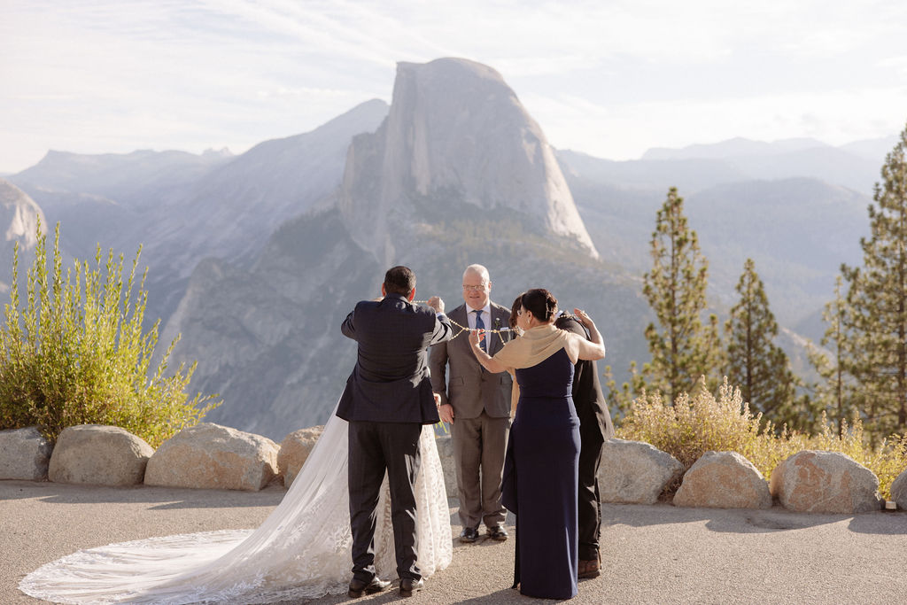 Bride and groom hold hands during an outdoor wedding ceremony with an officiant, set against a mountainous backdrop.
