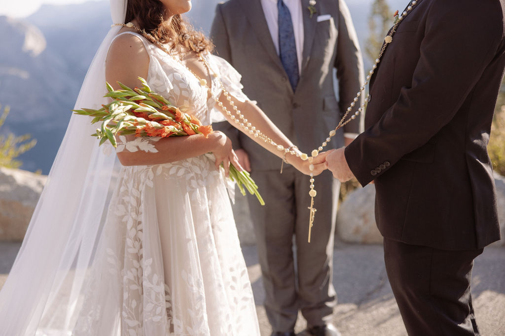 Bride and groom hold hands during an outdoor wedding ceremony with an officiant, set against a mountainous backdrop.