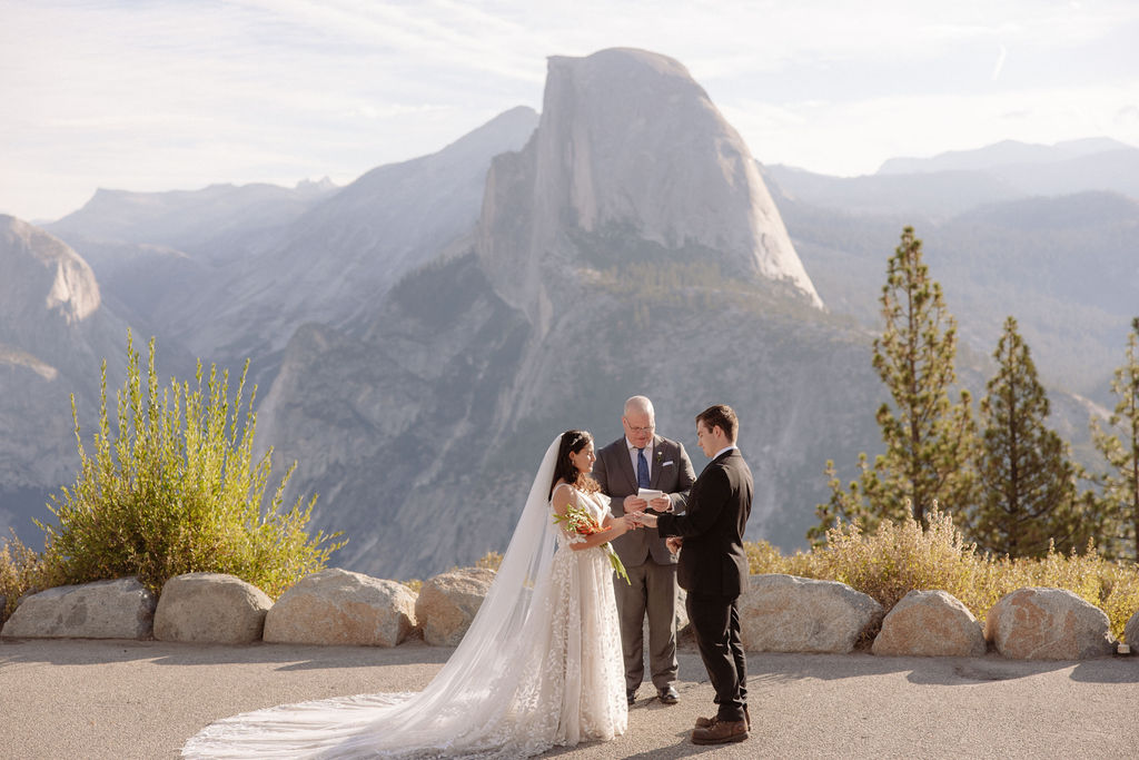 Bride and groom hold hands during an outdoor wedding ceremony with an officiant, set against a mountainous backdrop.