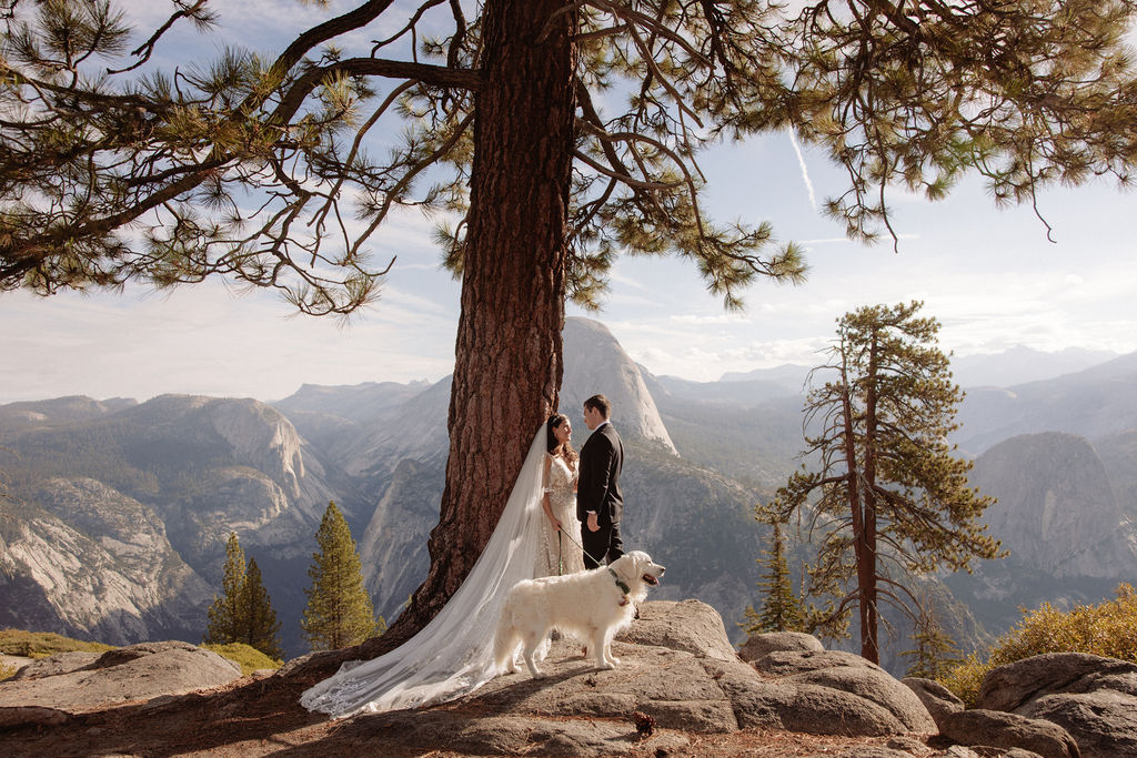 A bride and groom stand on rocky terrain overlooking a scenic mountain landscape at sunset, with the bride wearing a long white dress and veil for an elopement in Yosemite in September