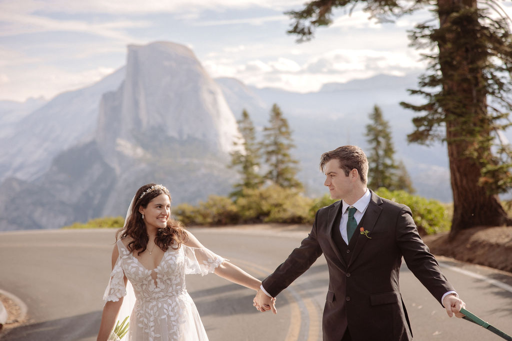 A bride and groom walk down a road lined with tall trees, accompanied by a white dog.