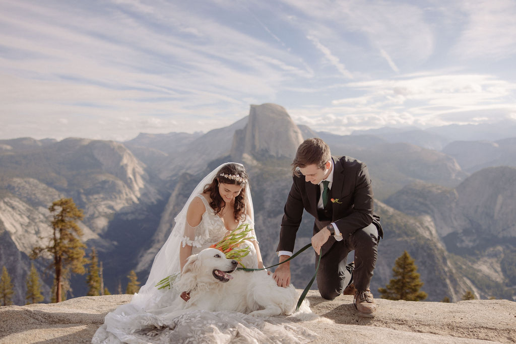 A bride and groom stand on a rocky ledge overlooking Yosemite Valley with Half Dome in the background under a partly cloudy sky for an elopement in Yosemite in September