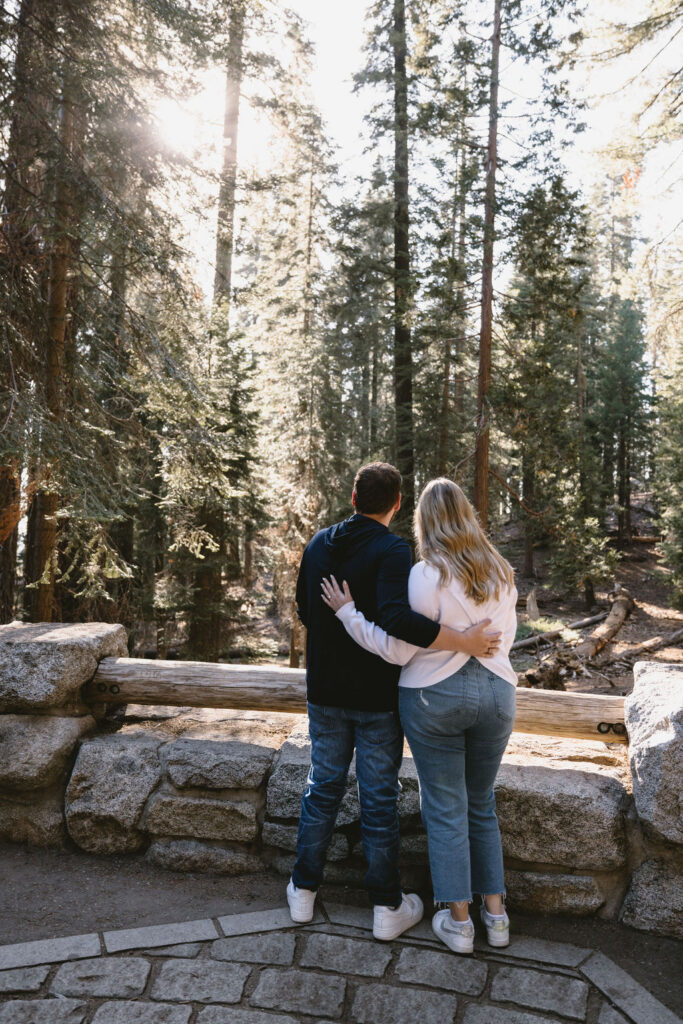 Two people stand side by side with arms around each other, facing a forest with tall trees and sunlight streaming through for Sequoia National Park Spots for Photos
