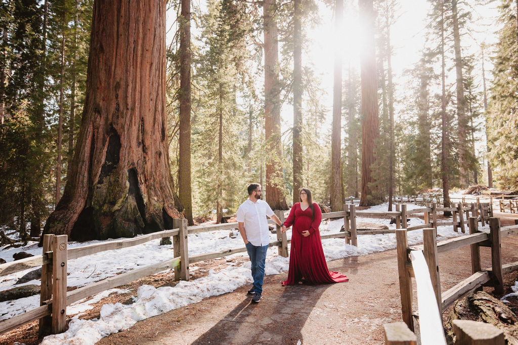 A couple stands close together outdoors near a wooden fence, with tall trees and sunlight in the background; the woman is wearing a long red dress.