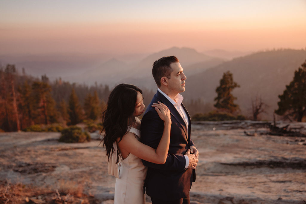 A woman in a light dress embraces a man in a suit from behind as they stand outdoors with mountains and trees in the background at sunset.
