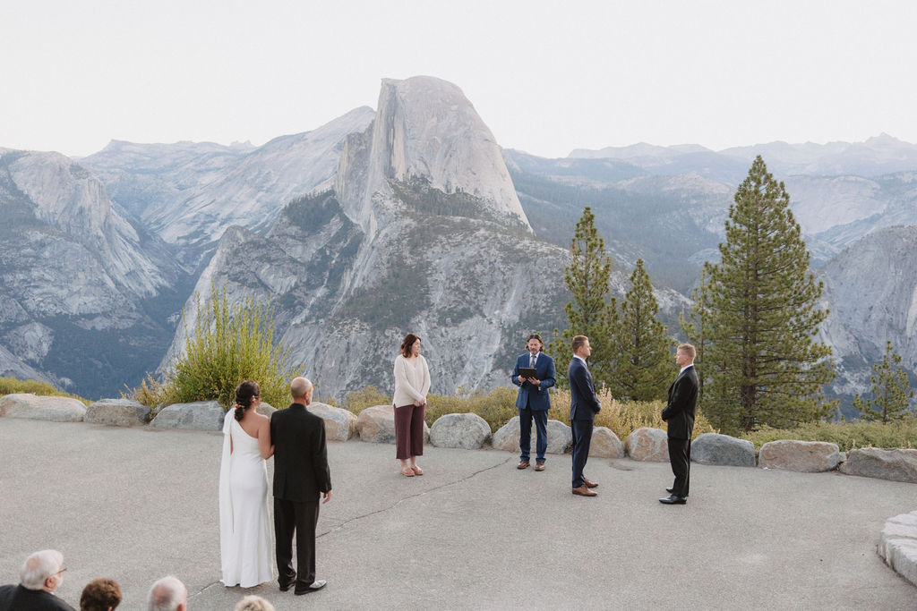 A wedding ceremony takes place outdoors with six people standing in a semicircle, overlooking a mountainous landscape with Half Dome in the background at Glacier Point Amphitheater