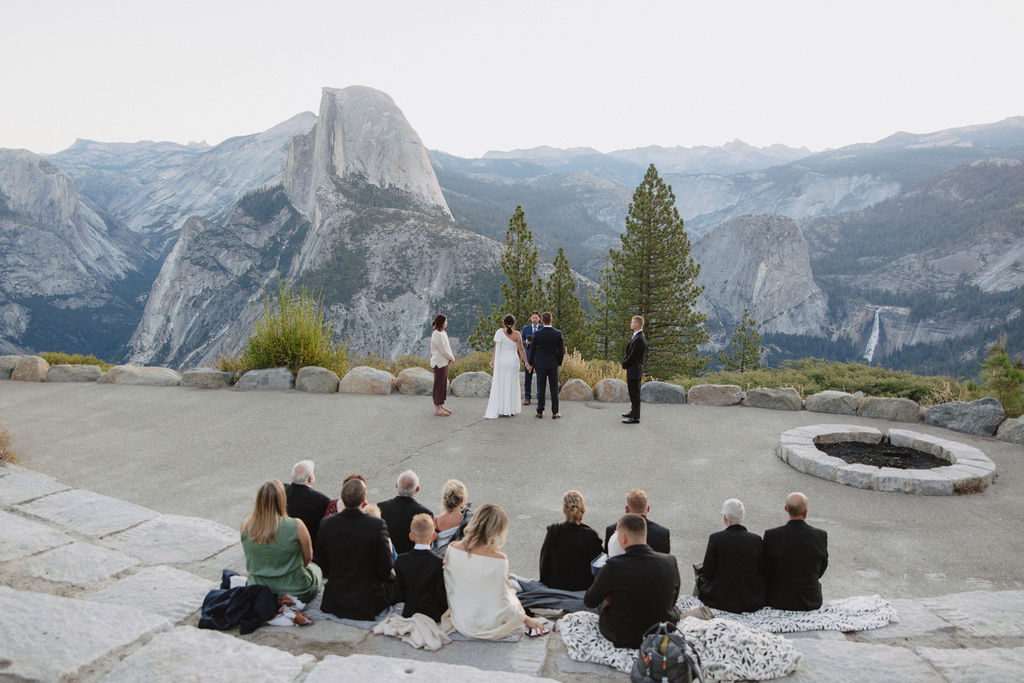 A wedding ceremony takes place outdoors with six people standing in a semicircle, overlooking a mountainous landscape with Half Dome in the background at Glacier Point Amphitheater