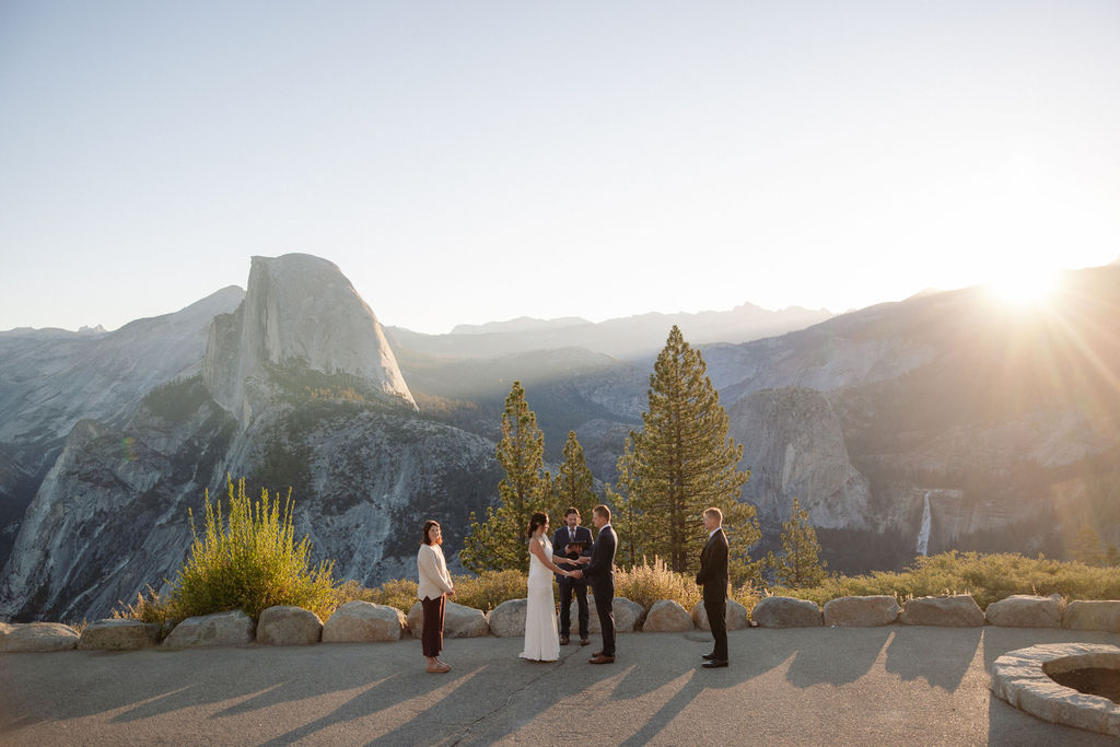A wedding ceremony takes place outdoors with six people standing in a semicircle, overlooking a mountainous landscape with Half Dome in the background at Glacier Point Amphitheater