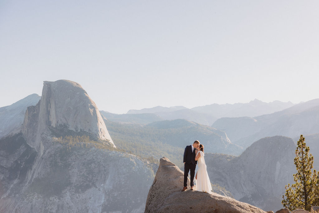 A bride and groom stand together on a rocky outcrop with mountain peaks, including Half Dome, visible in the background under a clear sky at Glacier Point Amphitheater