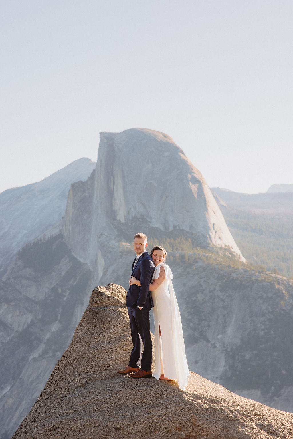 A bride and groom stand together on a rocky outcrop with mountain peaks, including Half Dome, visible in the background under a clear sky at Glacier Point Amphitheater