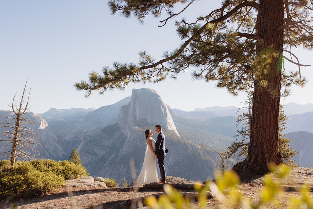 A couple dressed in wedding attire stands on a rocky ledge with mountainous scenery and a forested valley in the background.