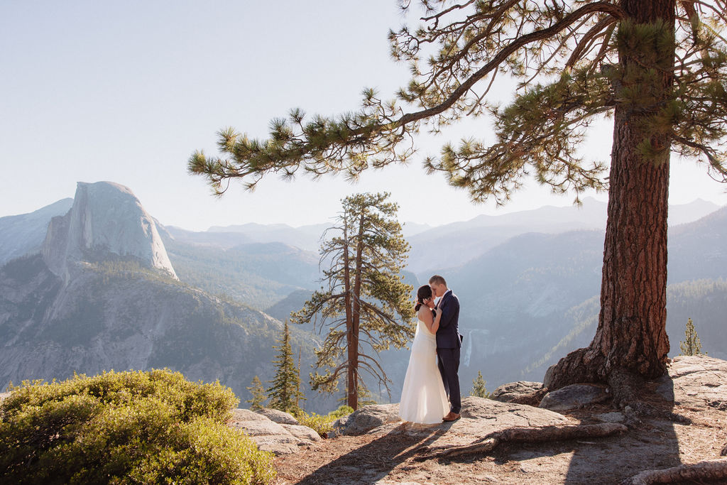A couple dressed in wedding attire stands on a rocky ledge with mountainous scenery and a forested valley in the background.
