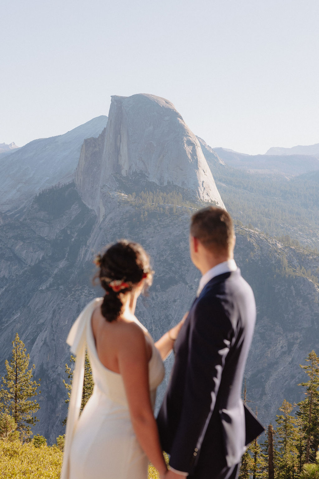 A bride and groom stand together on a rocky outcrop with mountain peaks, including Half Dome, visible in the background under a clear sky at Glacier Point Amphitheater