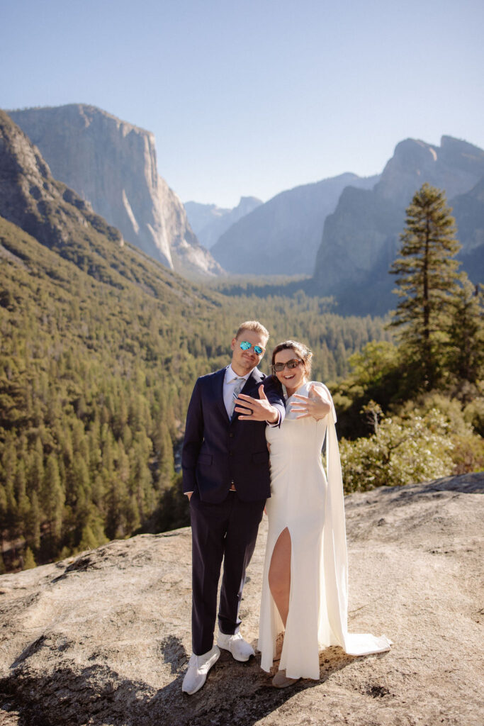 A couple dressed in wedding attire stands on a rocky ledge with mountainous scenery and a forested valley in the background.