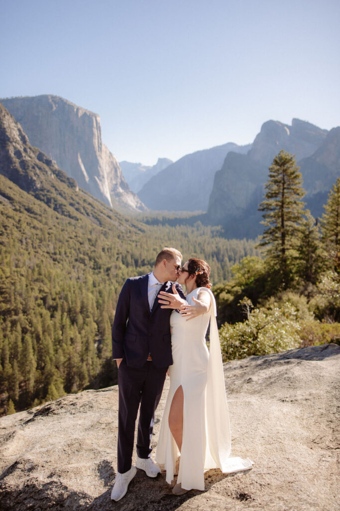 A couple dressed in wedding attire stands on a rocky ledge with mountainous scenery and a forested valley in the background.