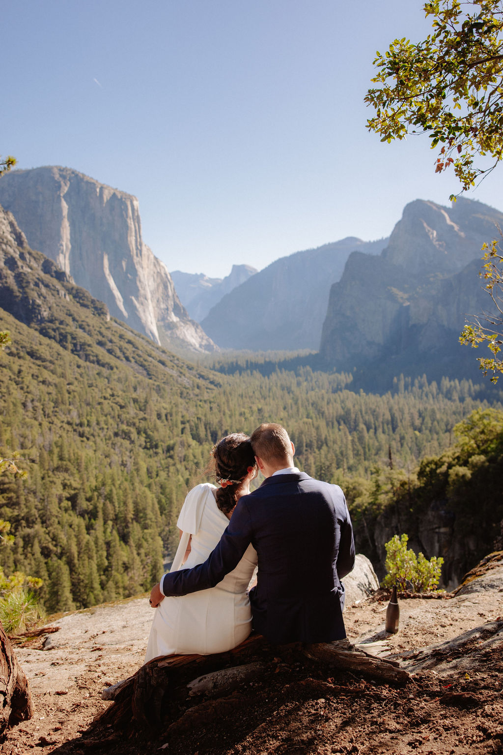 A bride and groom walk hand in hand on a wooden path through a field, with tall trees and rocky mountains in the background at Glacier Point Amphitheater