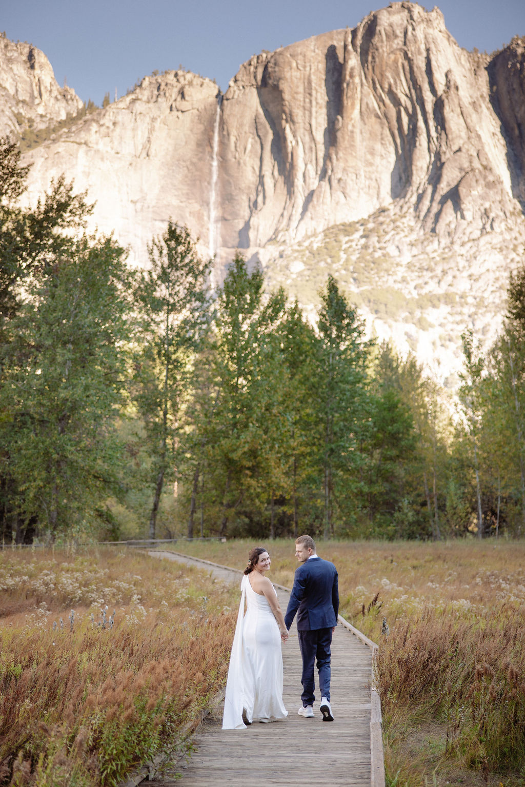 A bride and groom walk hand in hand on a wooden path through a field, with tall trees and rocky mountains in the background at Glacier Point Amphitheater