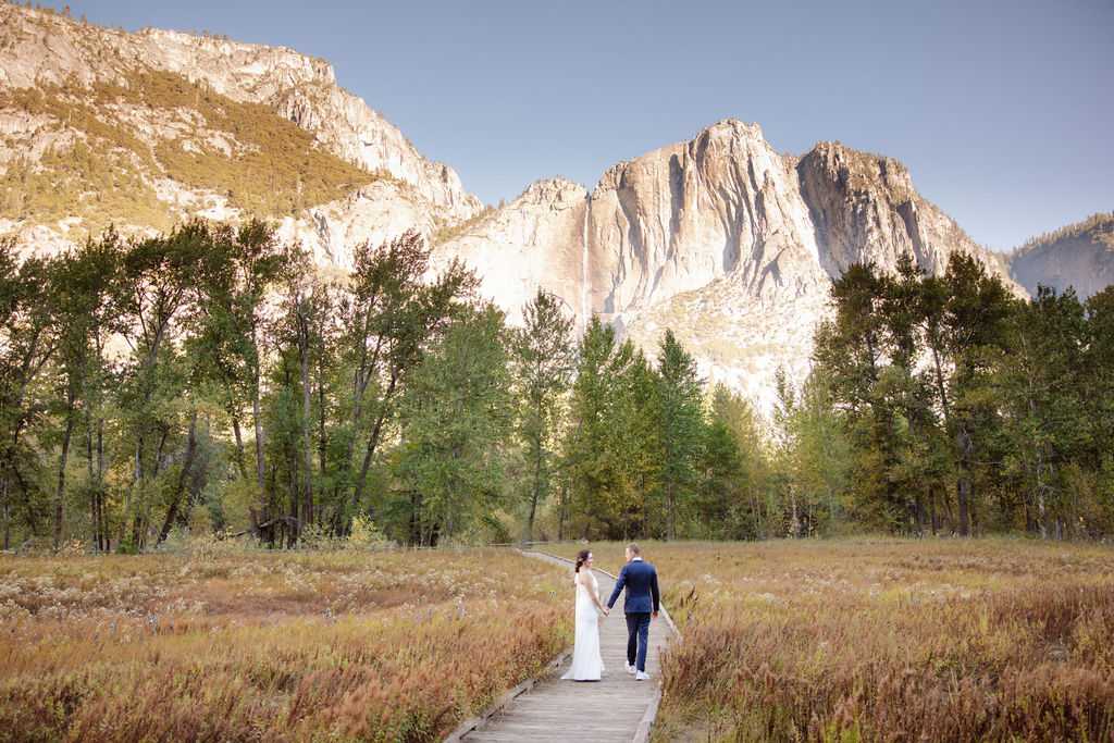 A bride and groom walk hand in hand on a wooden path through a field, with tall trees and rocky mountains in the background at Glacier Point Amphitheater