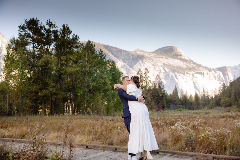 A bride and groom walk hand in hand on a wooden path through a field, with tall trees and rocky mountains in the background at Glacier Point Amphitheater