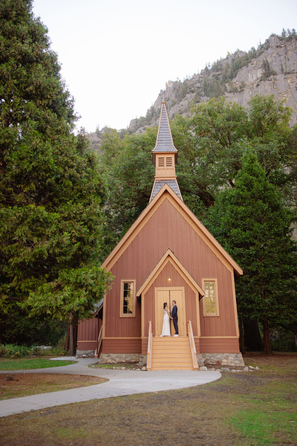 A bride and groom walk hand in hand on a wooden path through a field, with tall trees and rocky mountains in the background at Glacier Point Amphitheater