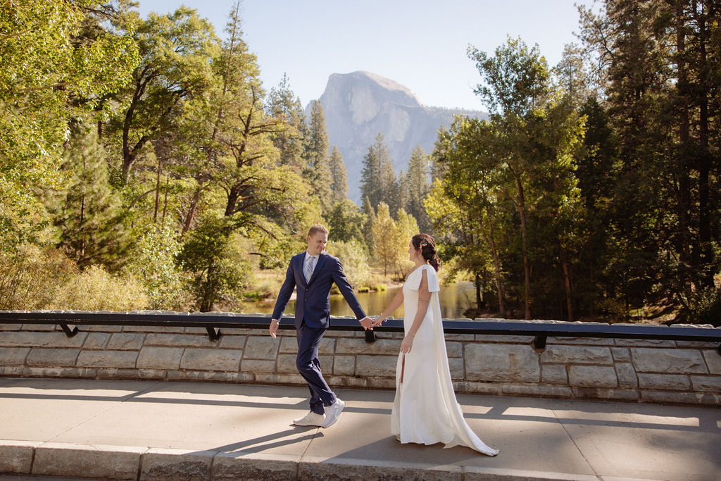 A couple dressed in wedding attire walks hand in hand on a bridge with trees and a mountain in the background.