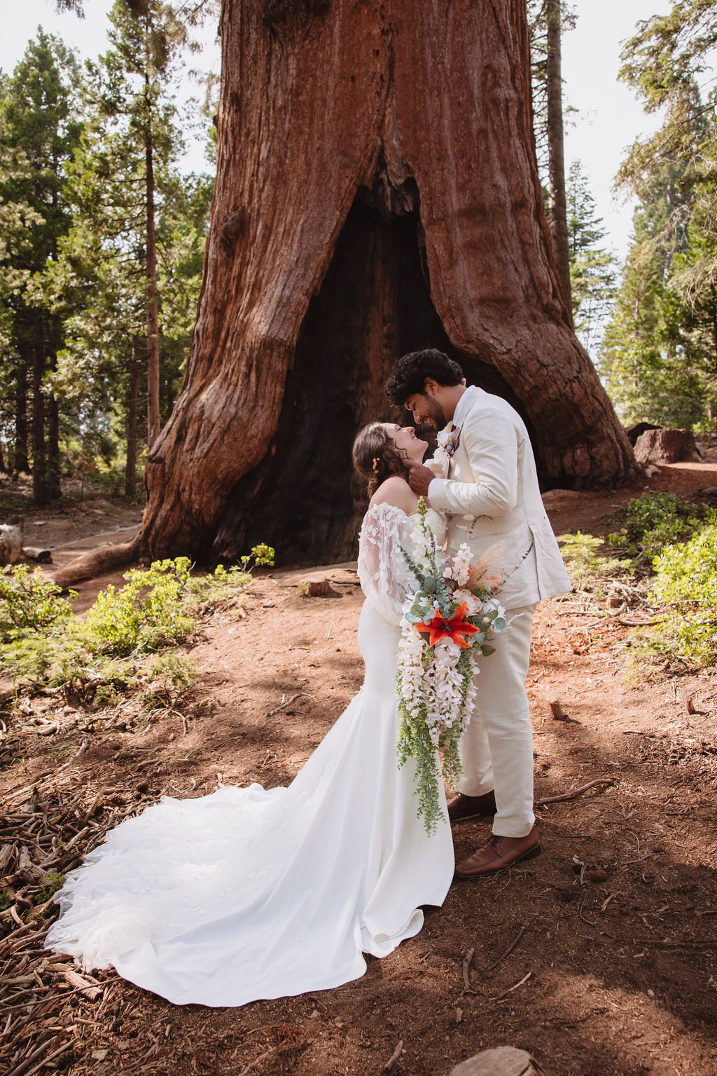 A bride and groom in wedding attire stand in front of a large tree in a forest, with sunlight filtering through the trees. Sequoia National Park Spots for Photos