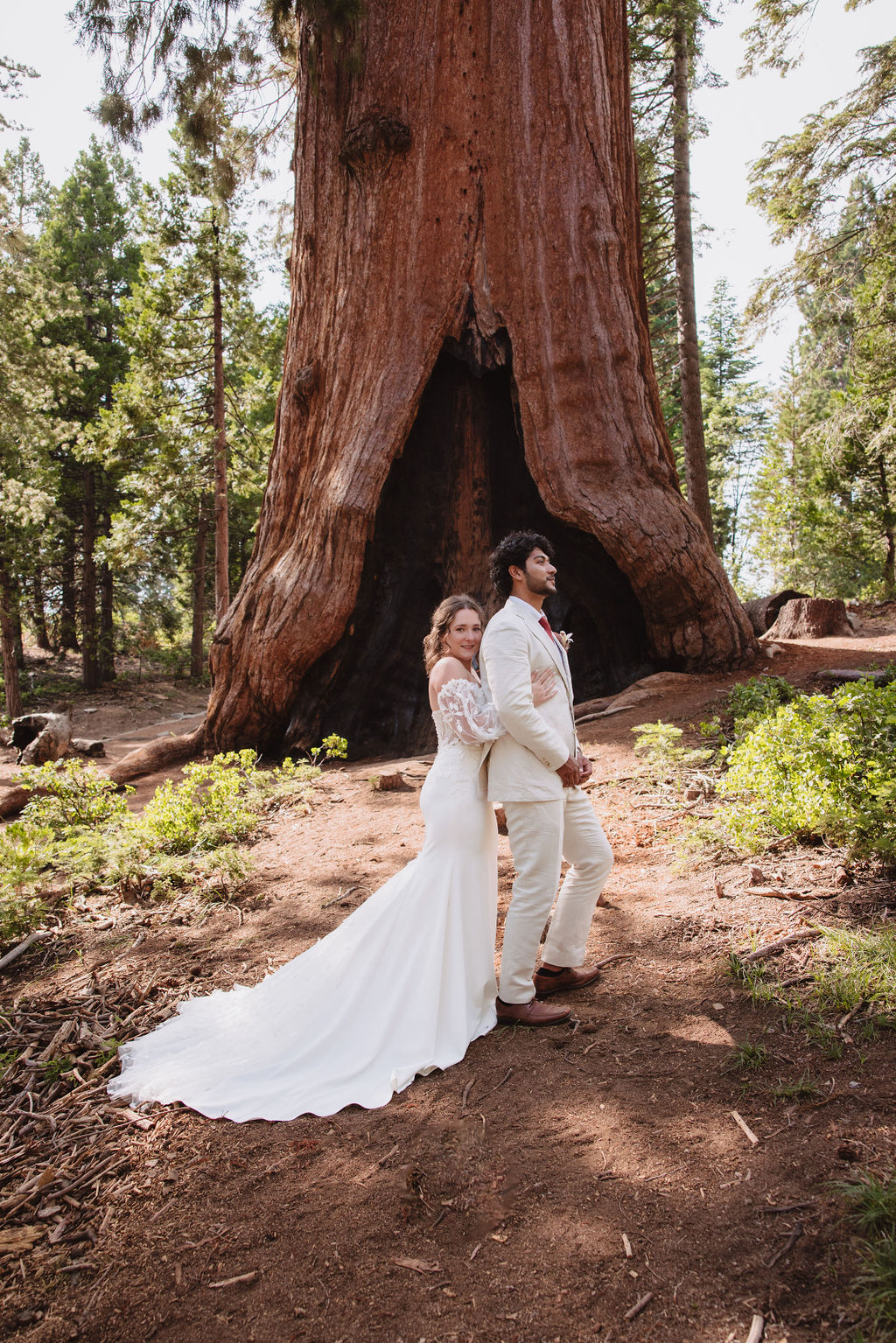 A bride and groom in wedding attire stand in front of a large tree in a forest, with sunlight filtering through the trees. Sequoia National Park Spots for Photos