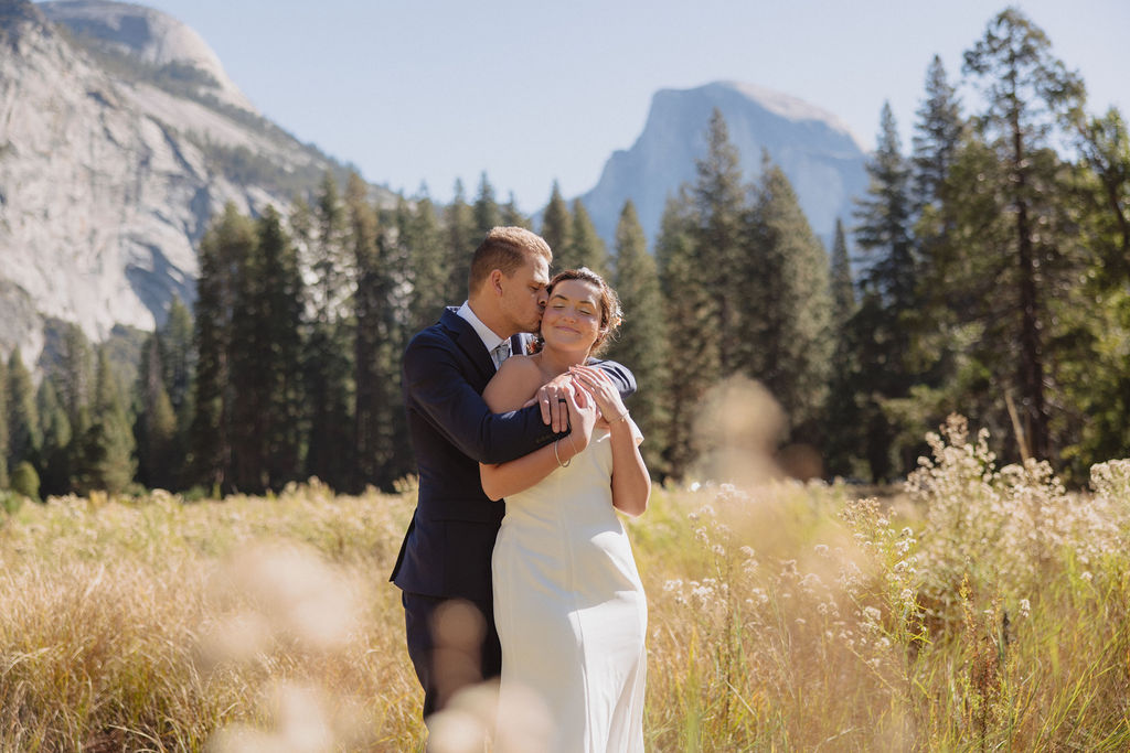 A bride and groom hold hands and look back at the camera while standing on a paved path, with mountains and trees in the background  at Glacier Point Amphitheater