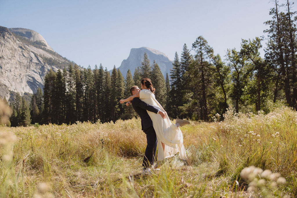 A bride and groom hold hands and look back at the camera while standing on a paved path, with mountains and trees in the background  at Glacier Point Amphitheater
