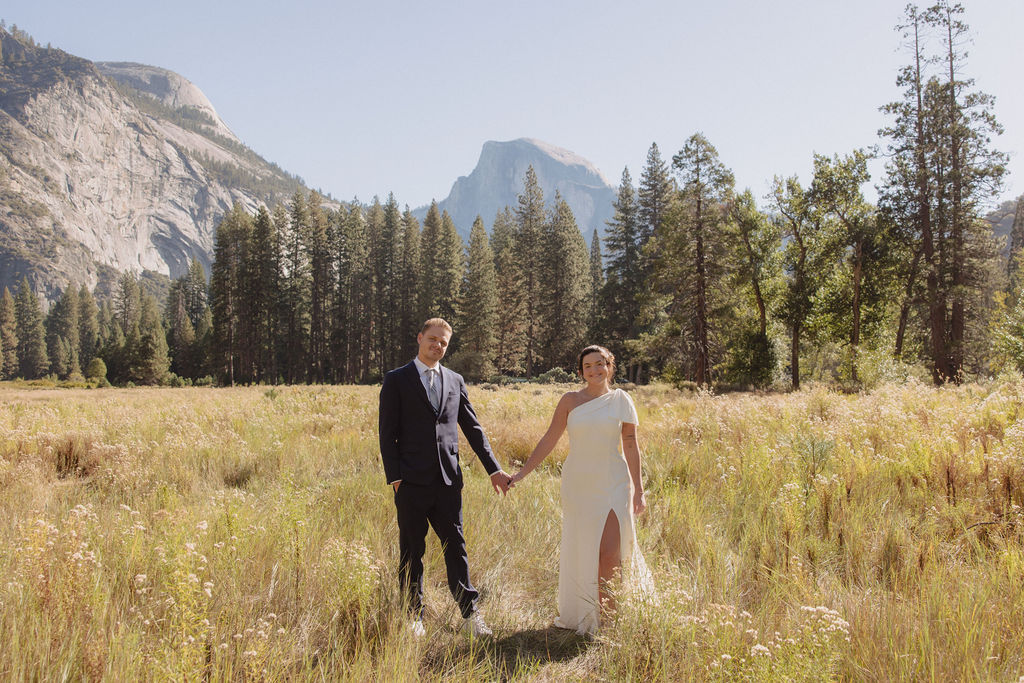 A bride and groom hold hands and look back at the camera while standing on a paved path, with mountains and trees in the background  at Glacier Point Amphitheater