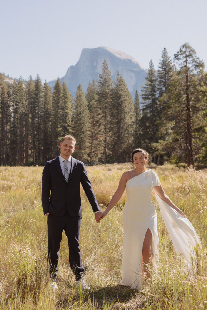 A bride and groom hold hands and look back at the camera while standing on a paved path, with mountains and trees in the background  at Glacier Point Amphitheater