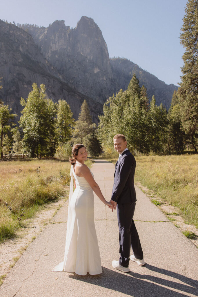 A bride and groom hold hands and look back at the camera while standing on a paved path, with mountains and trees in the background at Glacier Point Amphitheater